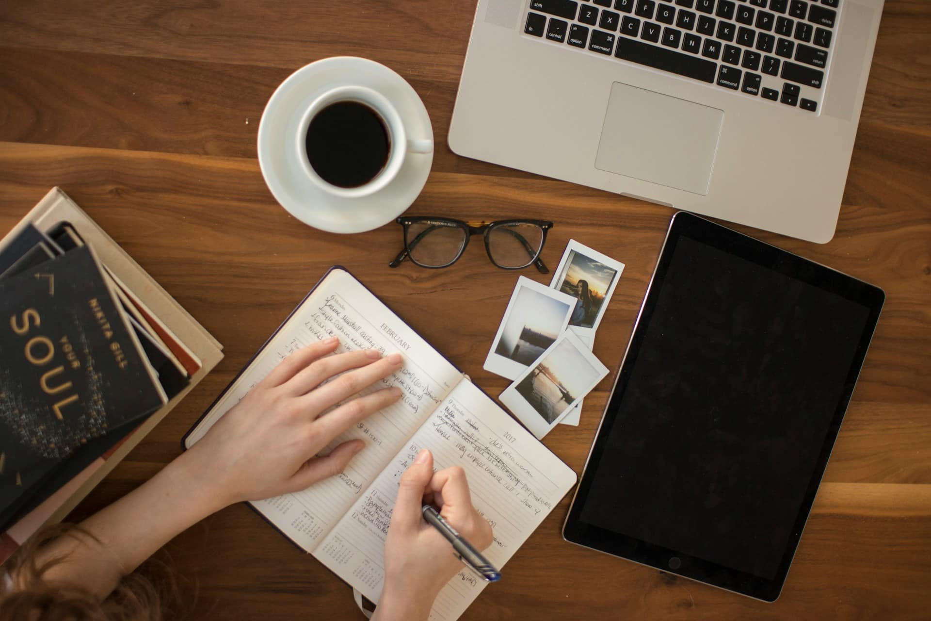 A person writing in a notebook at a desk beside a laptop and coffee